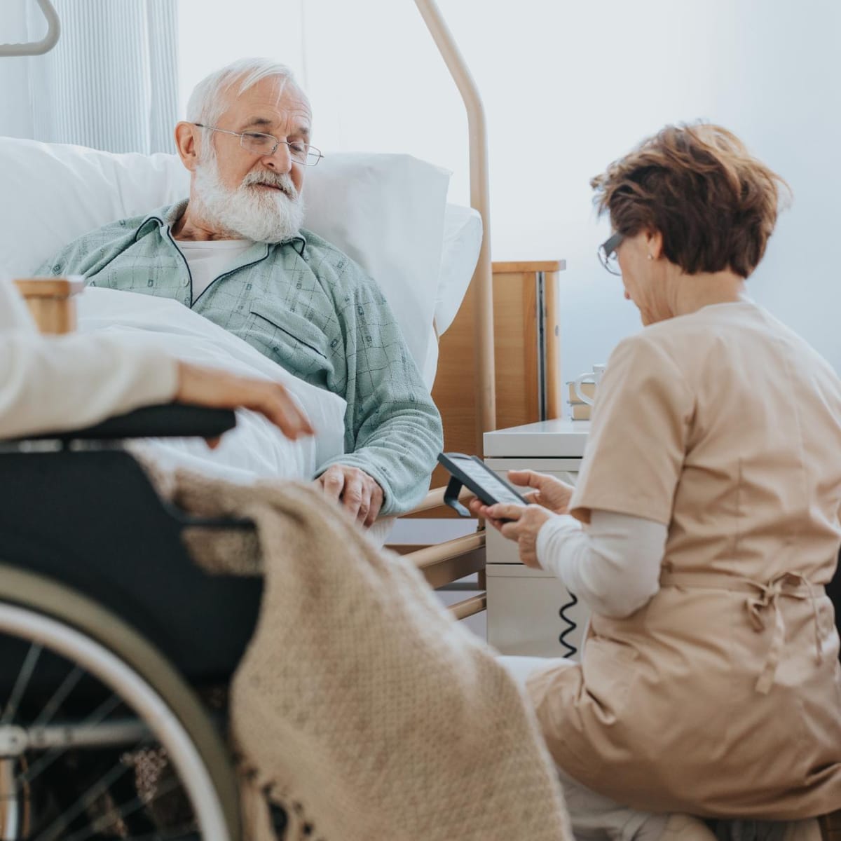 Caregiver kneeling down beside hospital bed asking an elderly man some questions